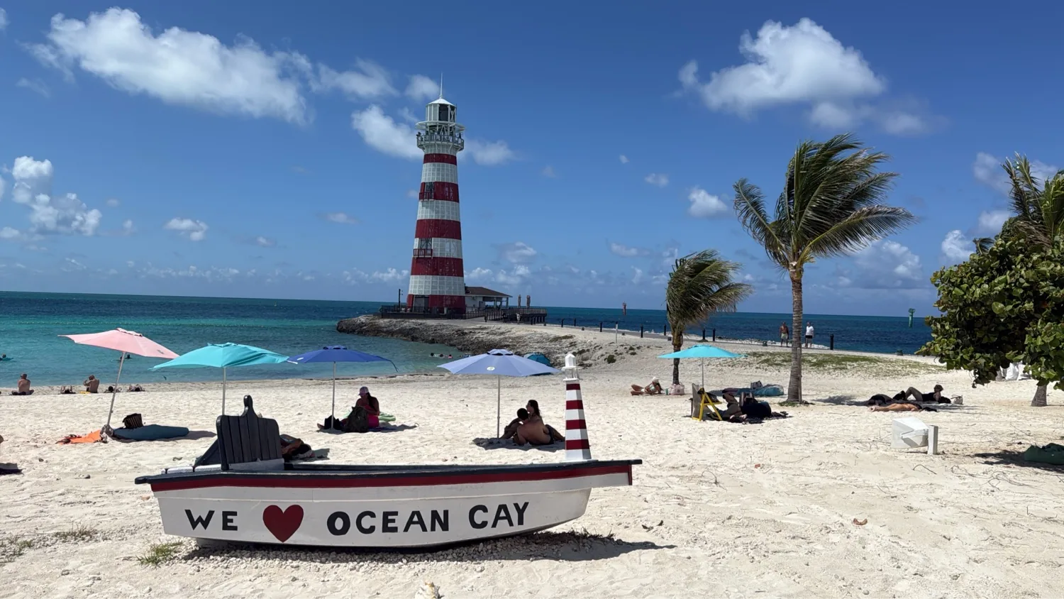 WE LOVE OCEAN CAY boat-bench on white sand with the red-and-white striped Marine Reserve lighthouse on a spit behind, beach umbrellas either side