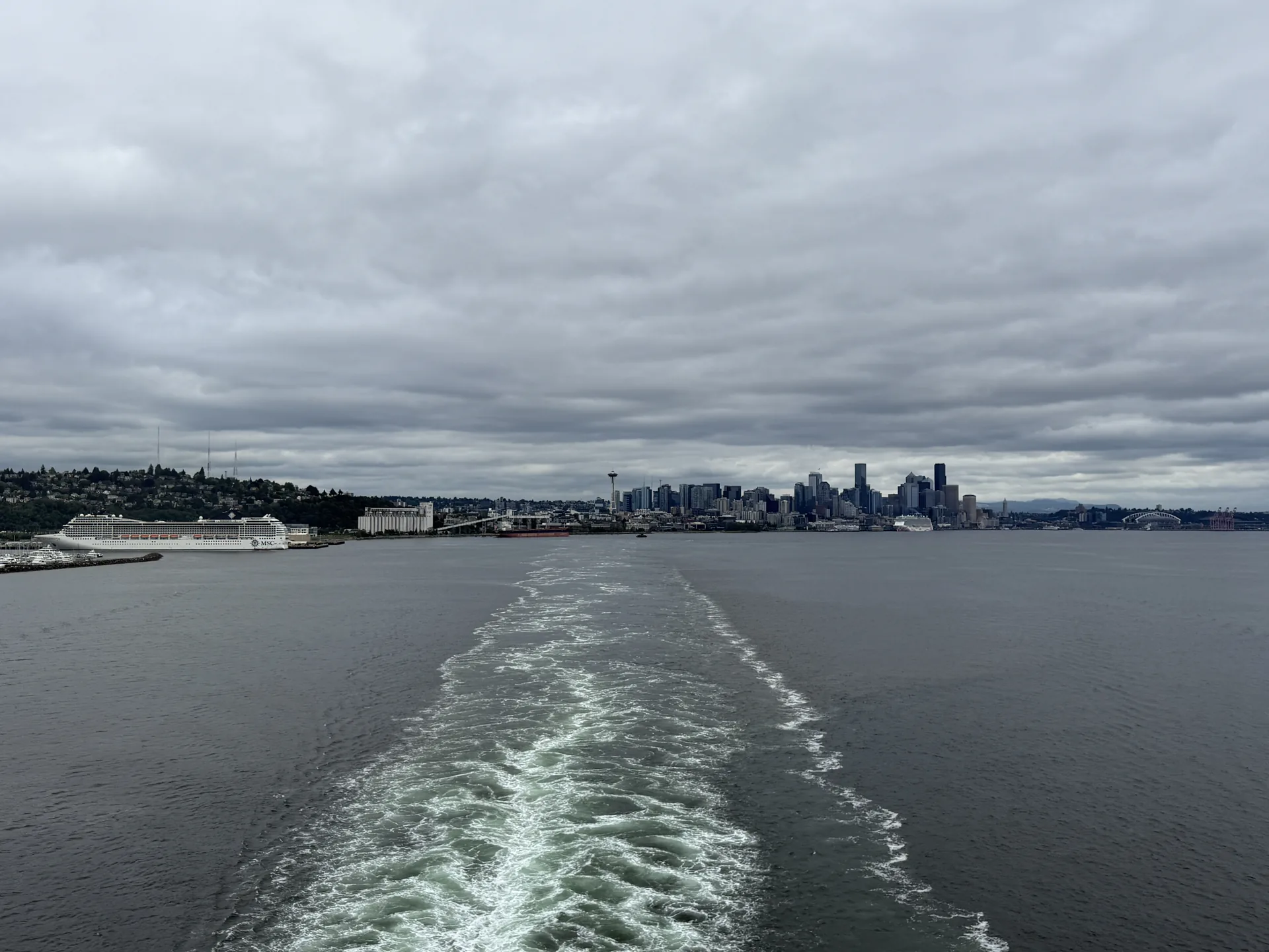 Departing Seattle by cruise ship: MSC Poesia berthed near the grain terminal at left, the downtown skyline and Space Needle at centre, and the ship's wake trailing across Elliott Bay