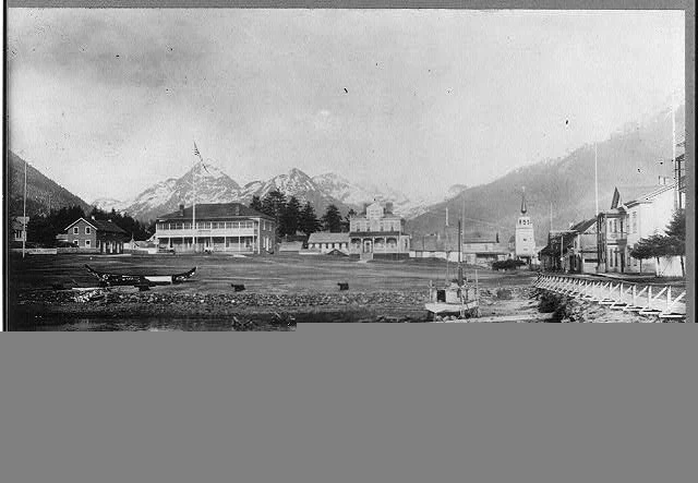 1906 panoramic black-and-white photograph of Sitka, Alaska from the parade ground with snow-capped mountains, cathedral, and waterfront buildings