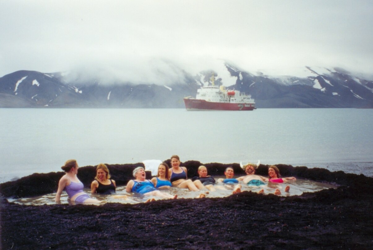 Tourists bathing in volcanic hot spring at Deception Island with expedition ship anchored in caldera behind them