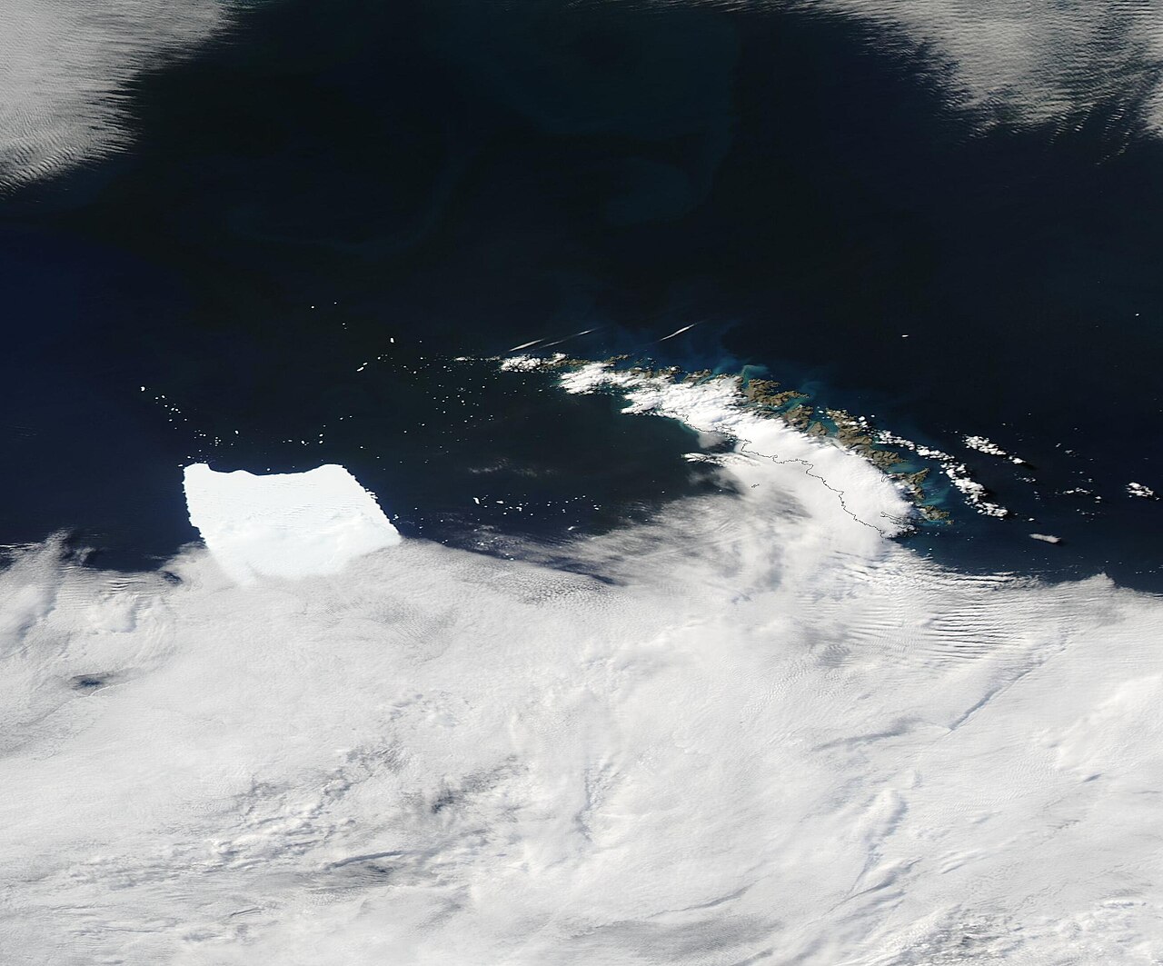 NASA MODIS satellite image showing the rectangular tabular iceberg A23a (white shape on left) in dark Southern Ocean water with the partially snow-covered South Shetland archipelago visible to the upper right under broken cloud cover