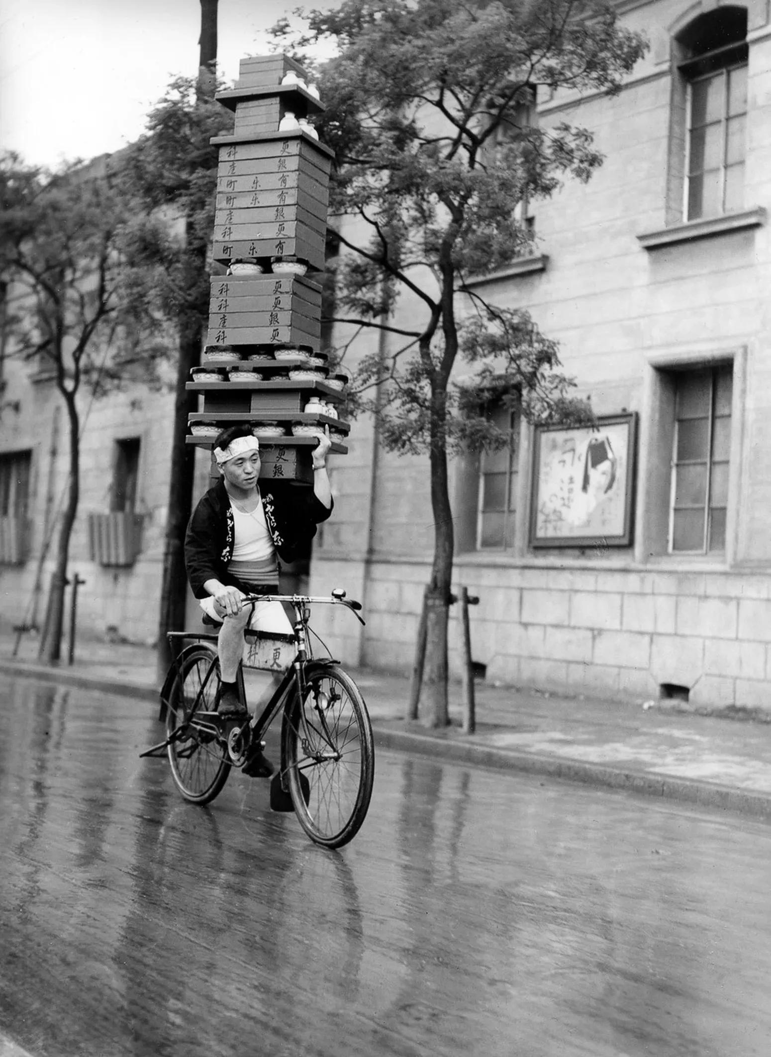 Historical black and white photograph of a Japanese soba delivery man cycling with tall stack of noodle boxes balanced on his head