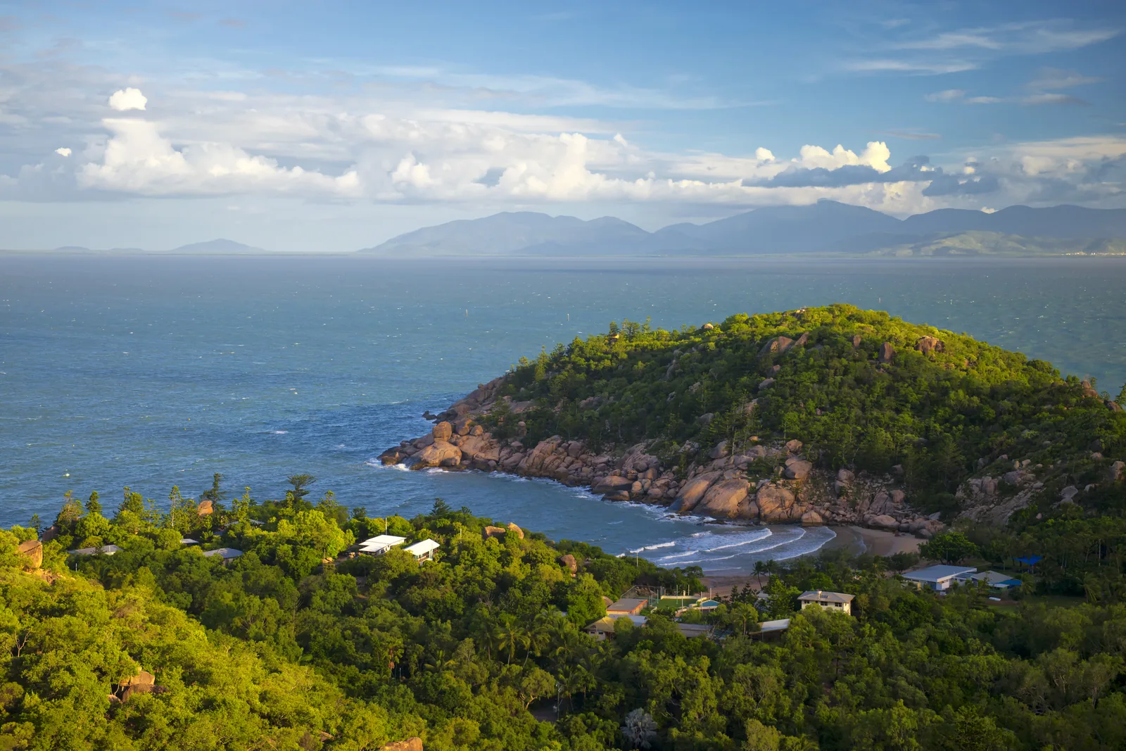 A golden-hour view from Magnetic Island over a granite-boulder headland and a small beach, with the mainland ranges blue across the channel
