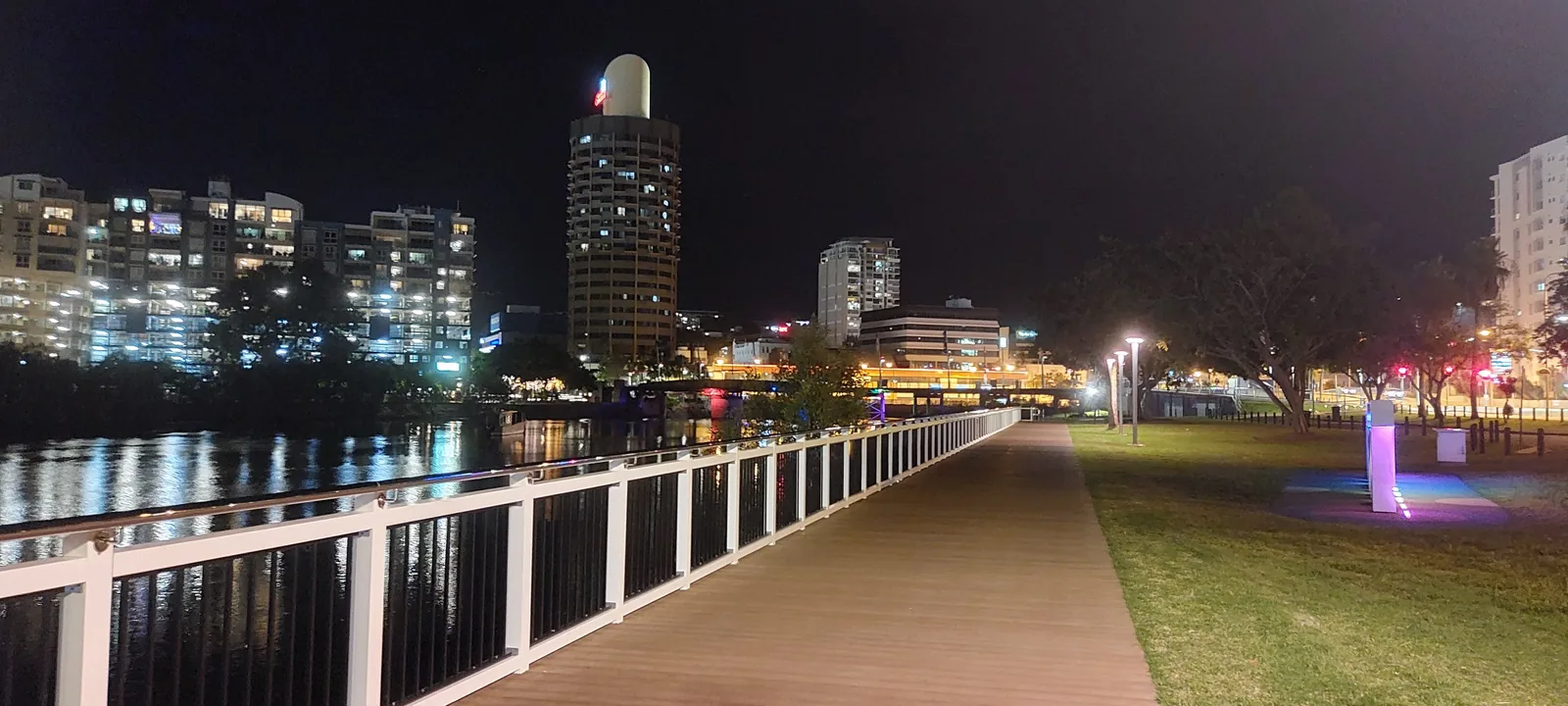 The Ross Creek boardwalk at night, its white railing leading toward the lit Townsville skyline including the round Sugar Shaker tower