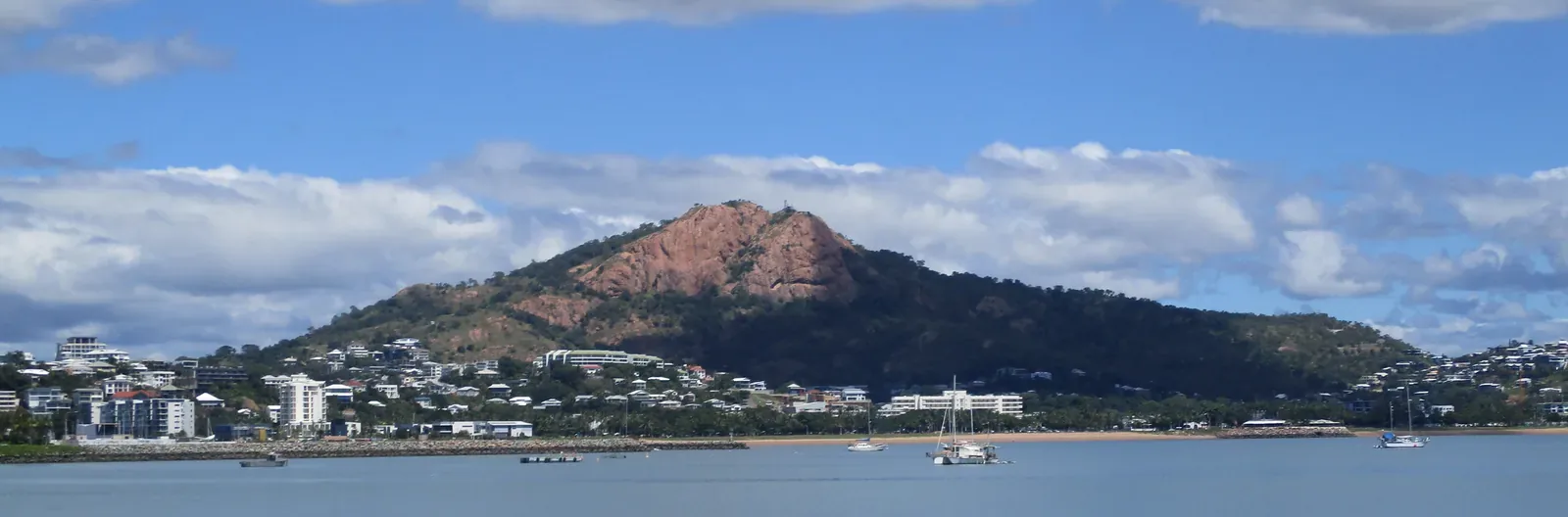 Castle Hill's pink granite dome rising above Townsville seen across the harbour, with the town along its foot, the breakwater and yachts at anchor