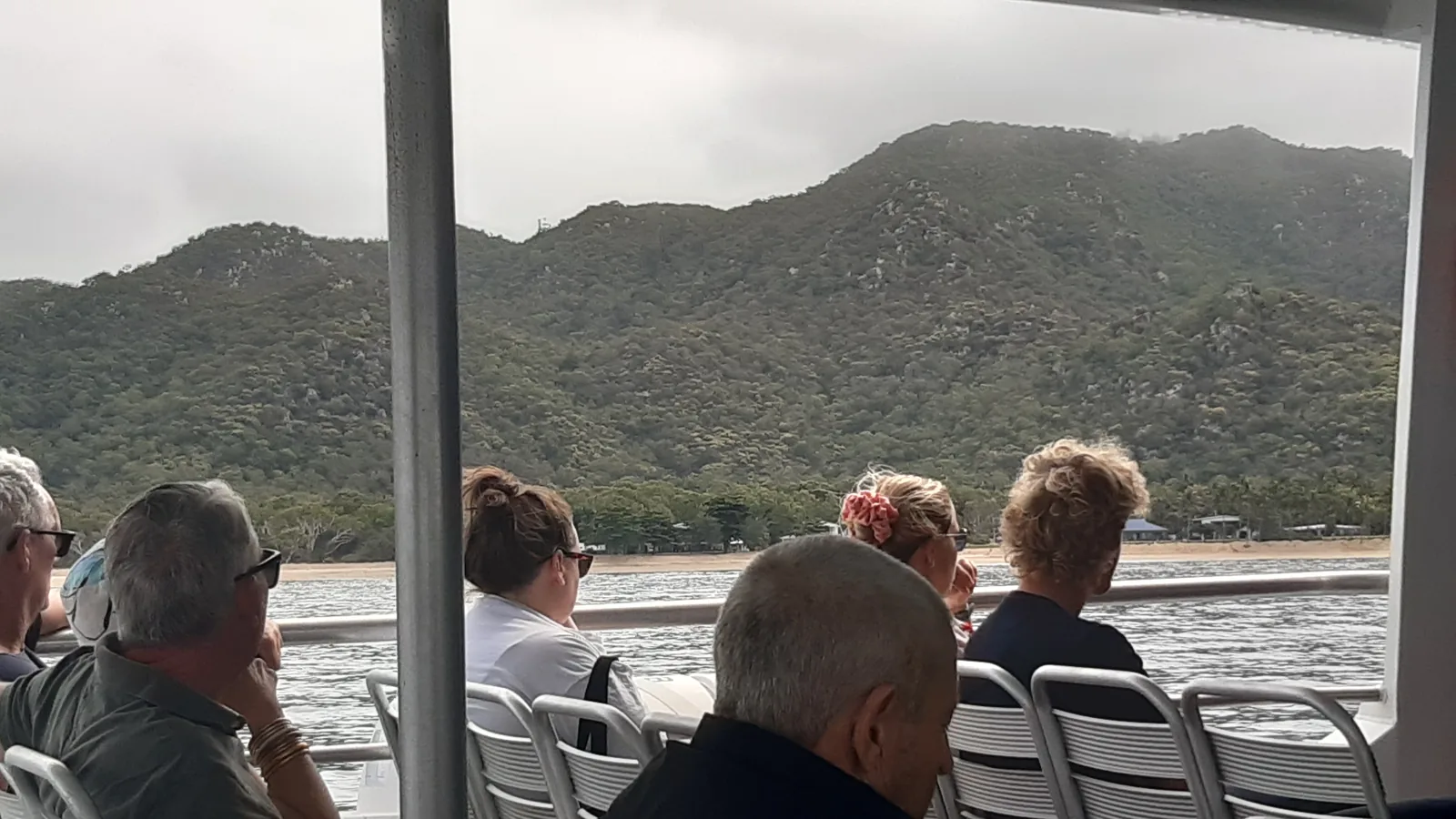 Seated ferry passengers seen from behind under an awning as the hills of Magnetic Island come up ahead across grey water