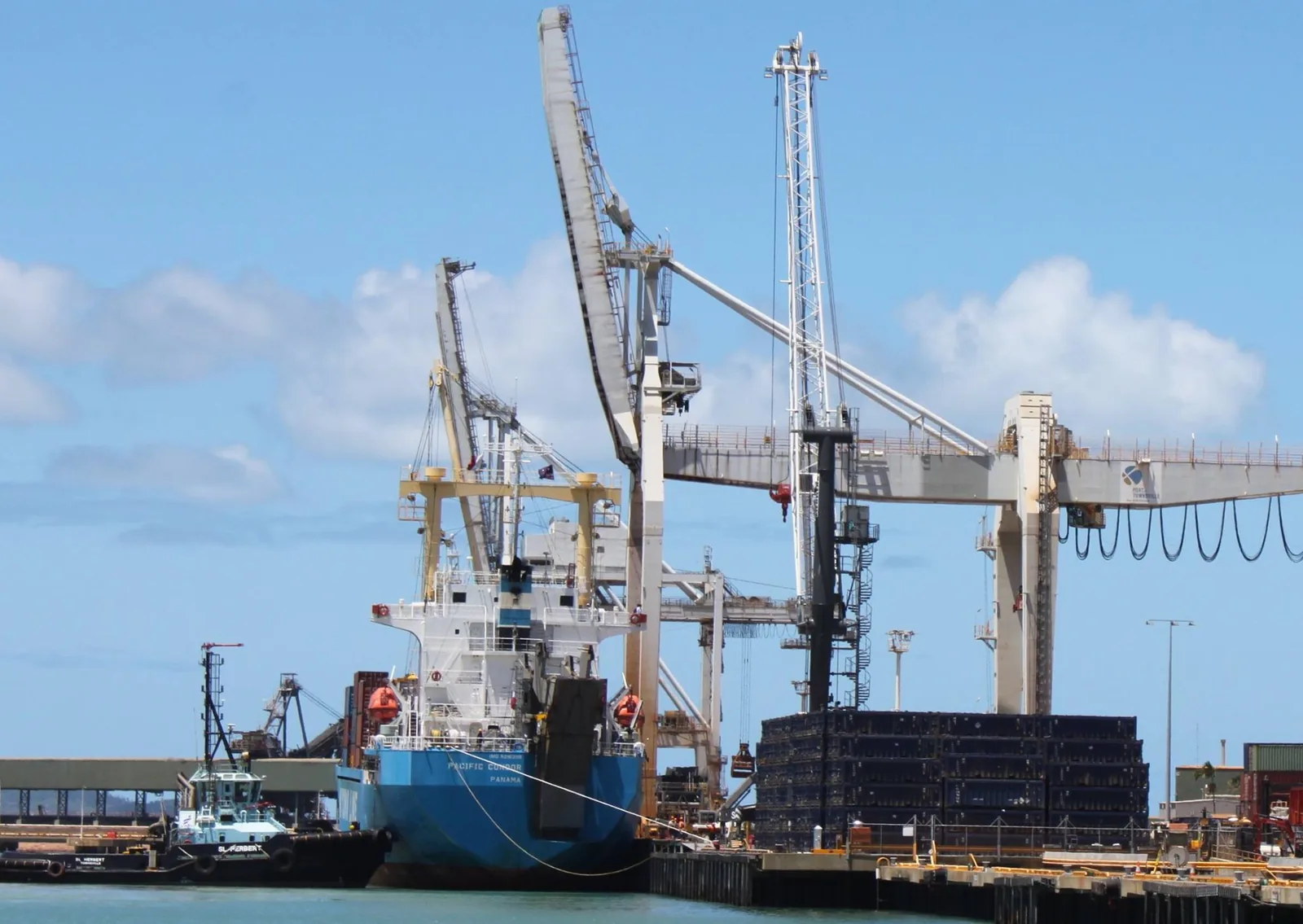 A general cargo ship alongside a quay at the Port of Townsville with its cranes raised, a shore gantry over stacked cargo and a tug at left