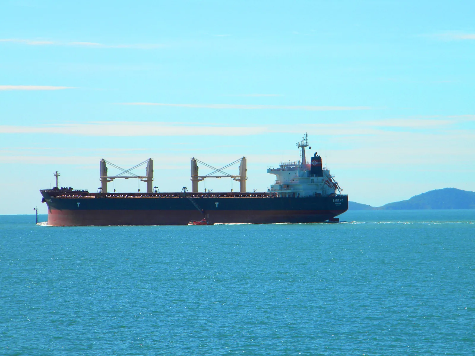 A bulk carrier under way in open water with a pilot boat alongside her hull and low hills behind