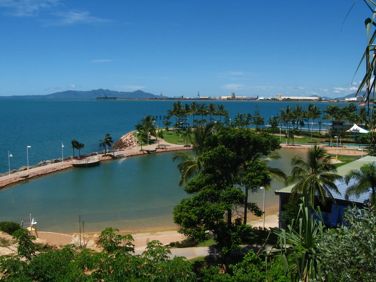 An elevated view over the Strand rock pool at Townsville — a stone-edged saltwater lagoon with palms, a waterpark, the port jetty and the Cape Cleveland ranges across the bay