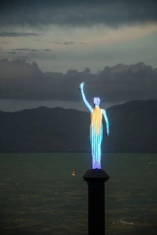Ocean Siren, an illuminated figure sculpture standing on a pile at the water's edge at dusk with one arm raised holding a shell