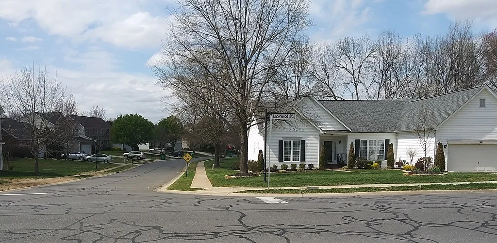 A quiet American suburban street corner with wide lanes and driveways—symbol of car-centric travel.