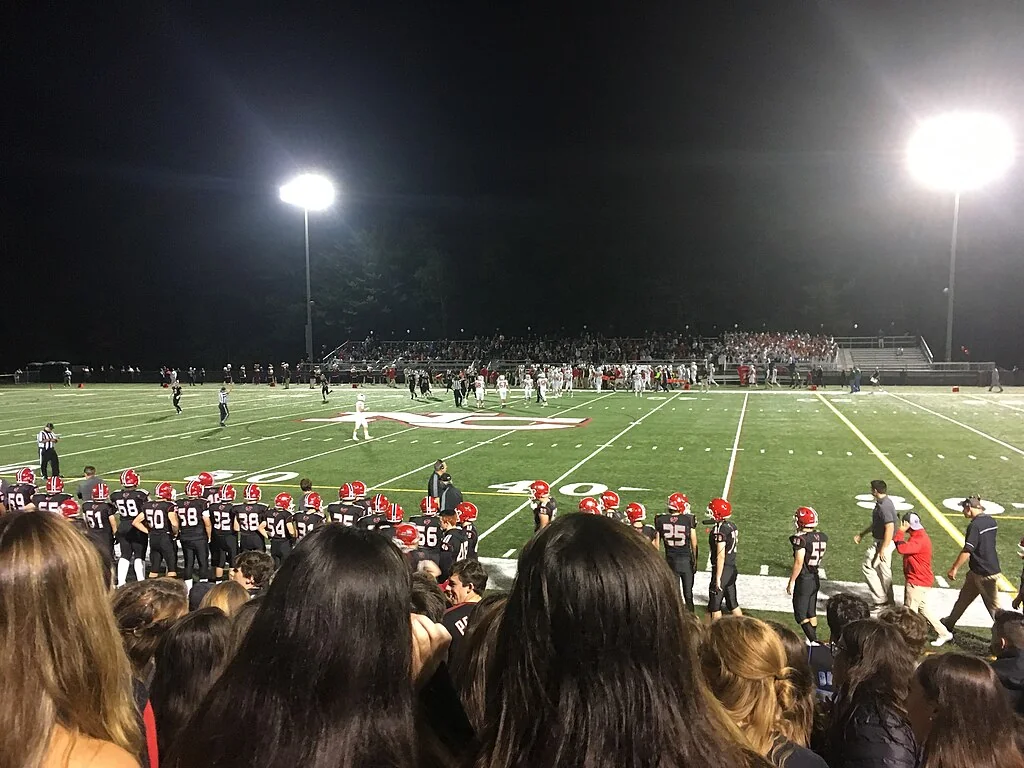 Friday-night lights at a U.S. high school football game — school-spirit culture in full color.